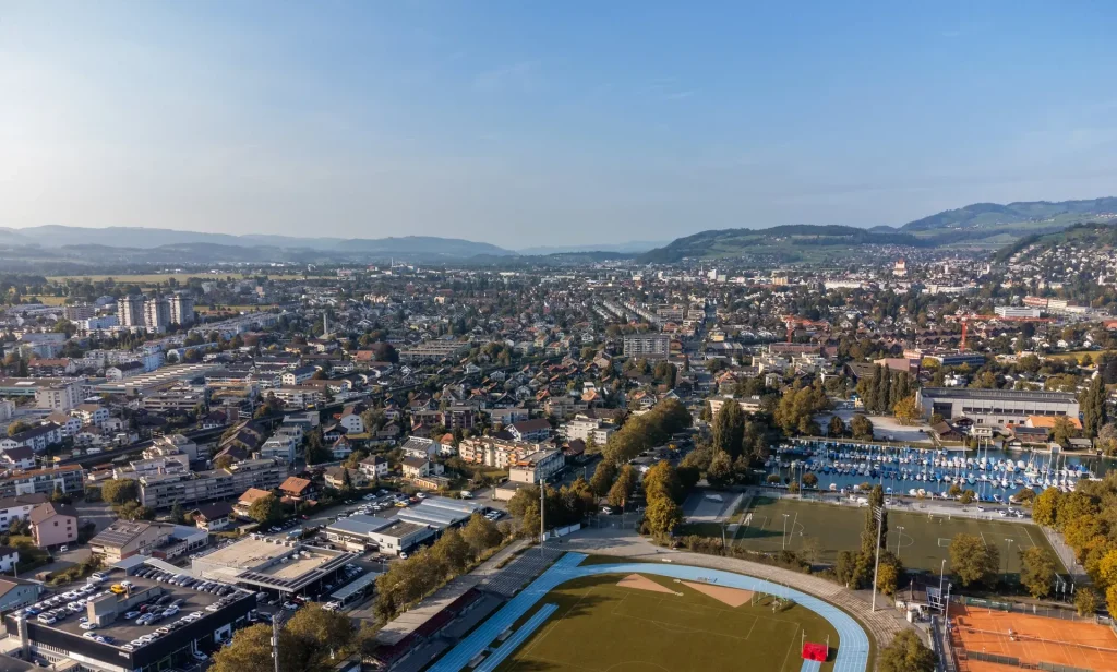 Das Dürrenast von oben: Häuser, Sportplätze und ein Bootshafen am See, dahinter die Stadt Thun.